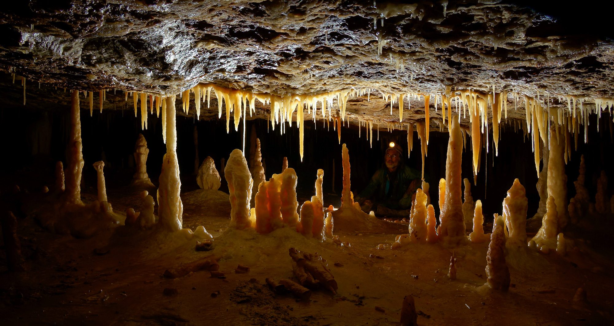 Stalactites and stalagmites glowing orange in backlight; a caver in the background admires the scene.